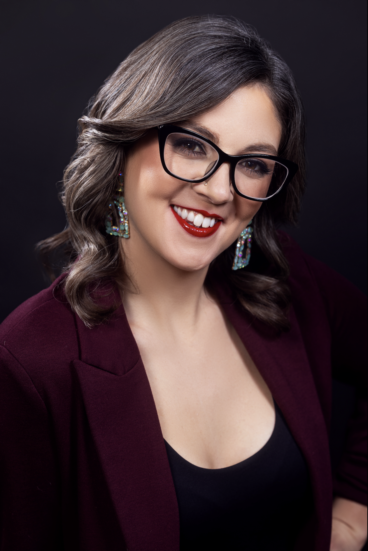 Alyssa Allgood wears a blazer, glasses, and a pair of jewel earrings. She smiles and and stands in front of a black backdrop.